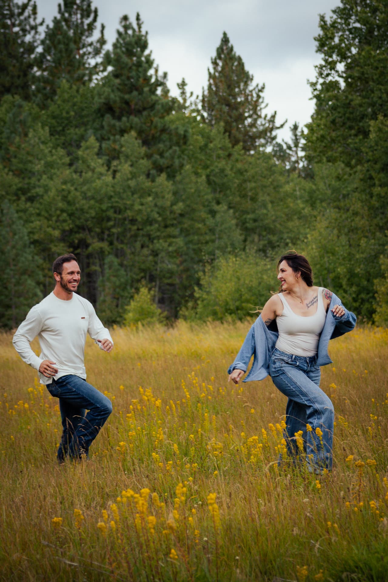 Couple laughing and dancing together in a golden wildflower meadow with pine trees behind them