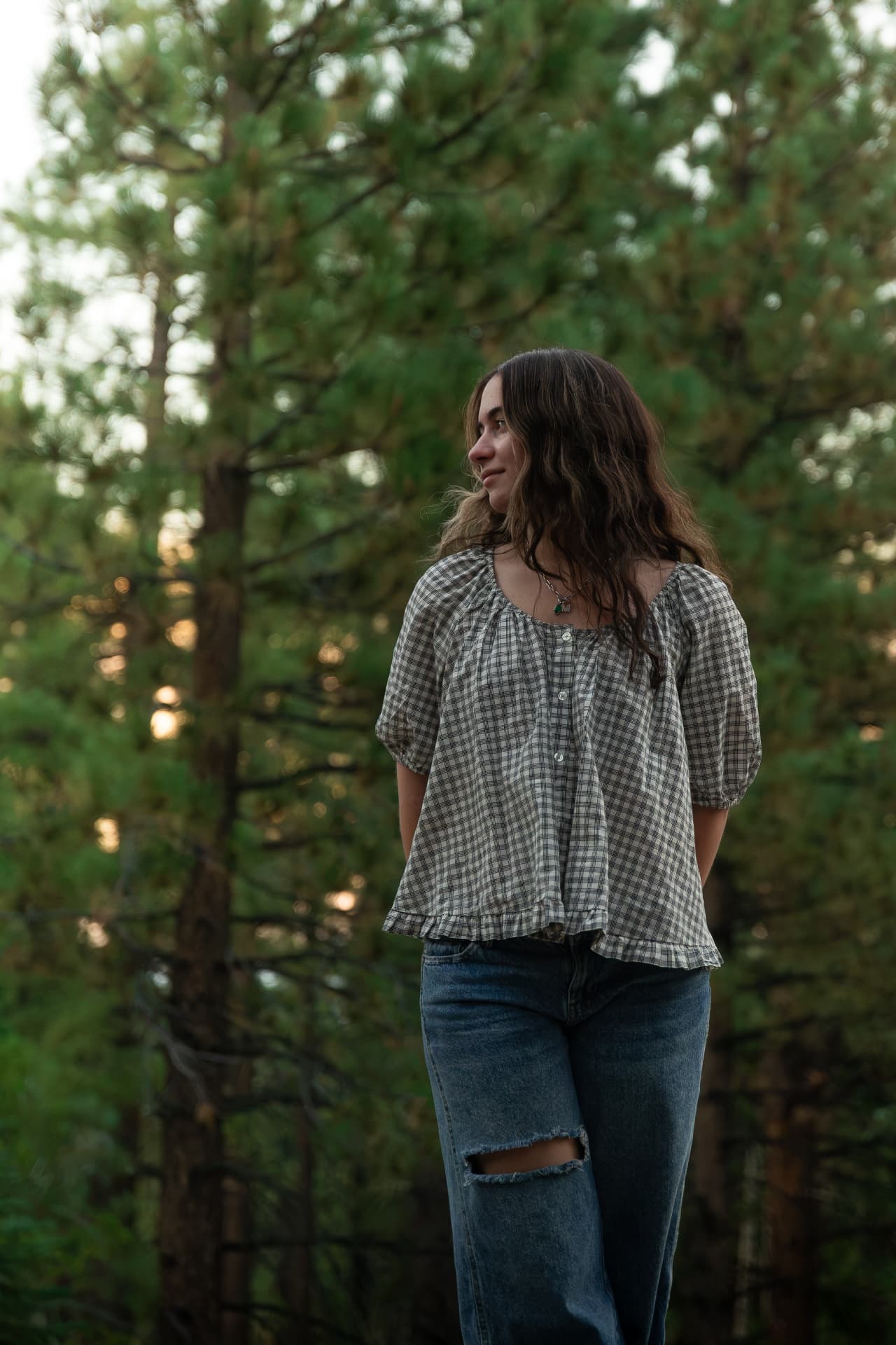 Senior portrait of a young woman in a plaid blouse looking off into tall backlit pines