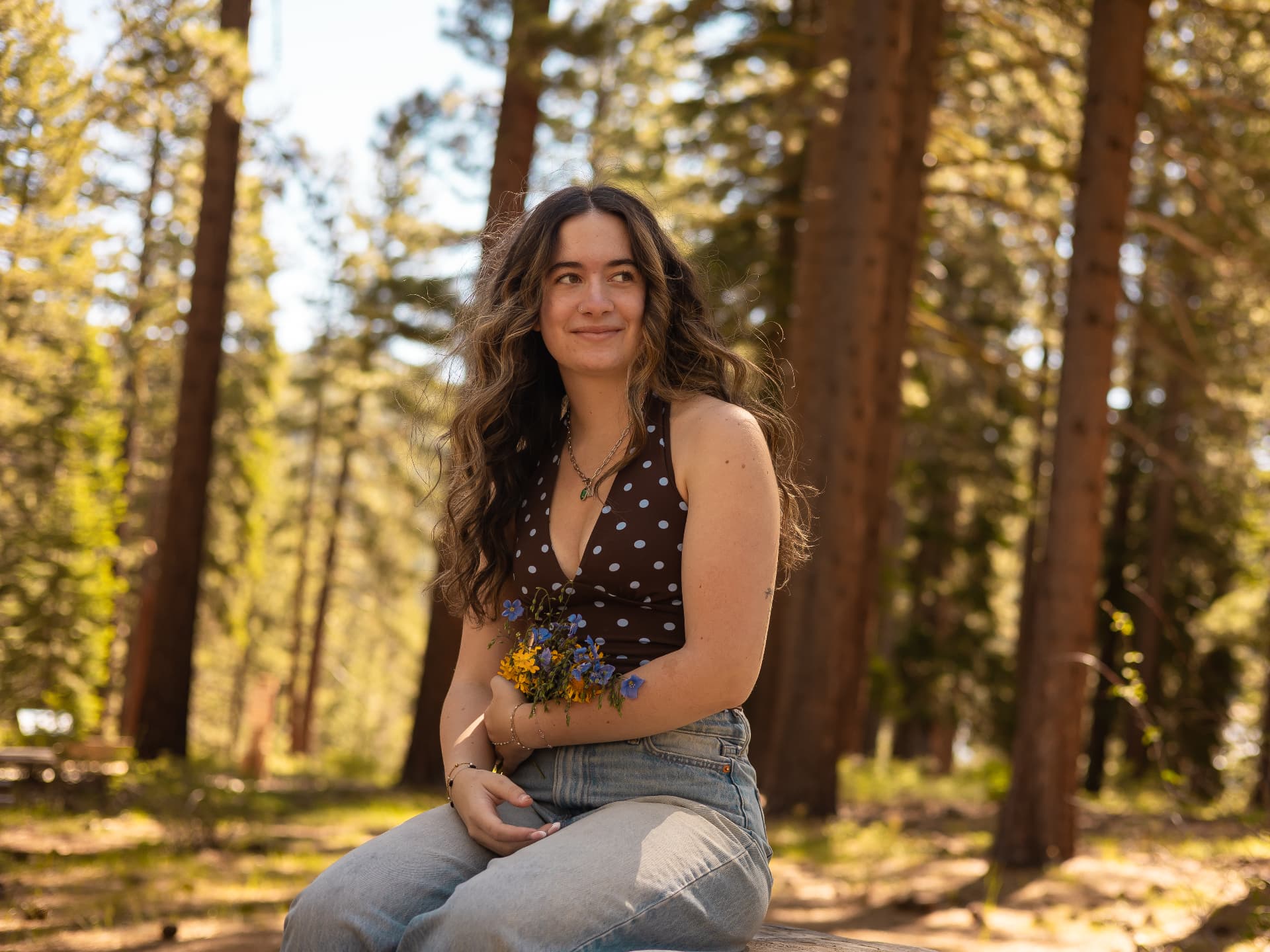 Senior portrait of a young woman sitting on a log with a handful of wildflowers among tall pines