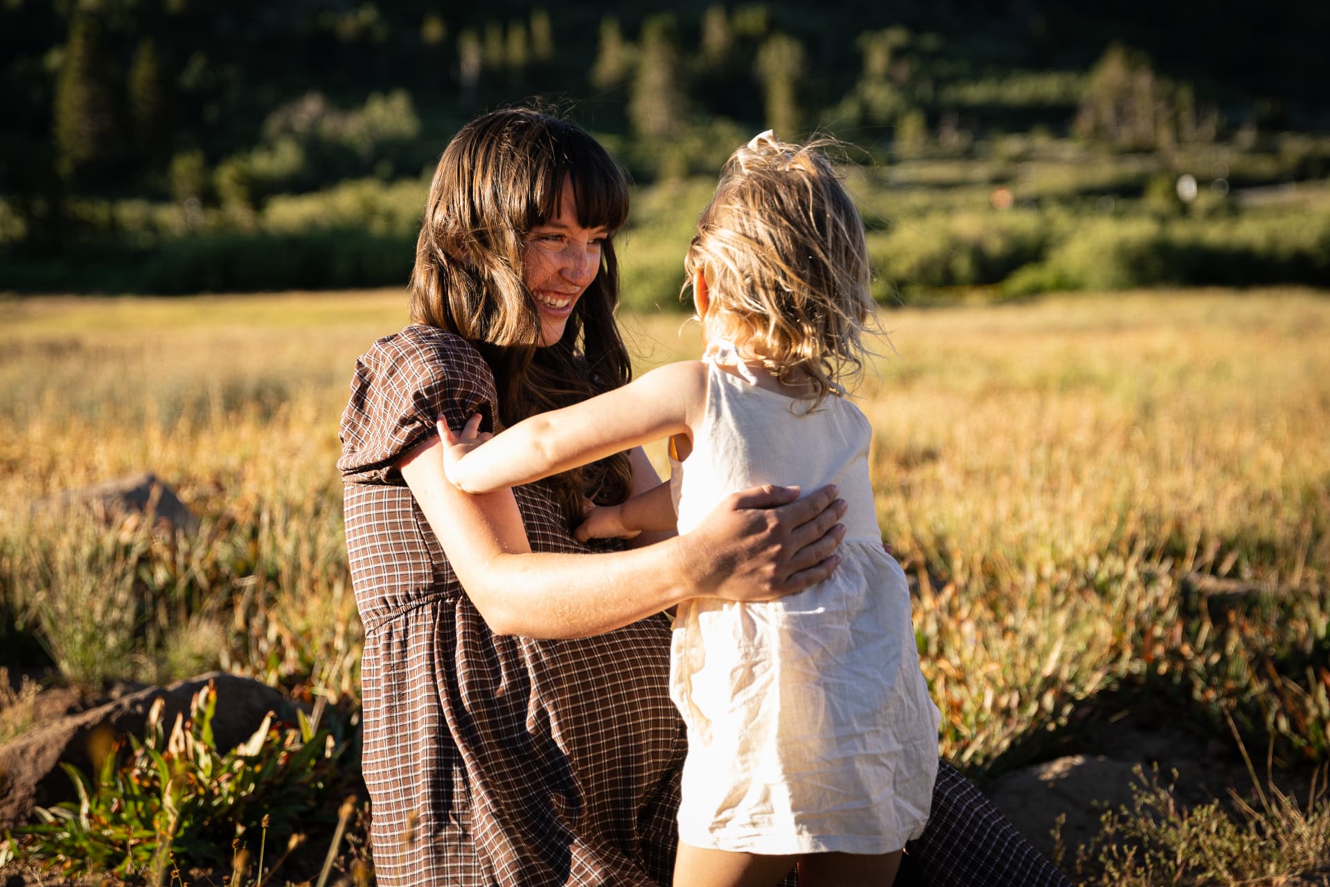 Mother laughing as her young daughter hugs her in a golden mountain meadow at sunset