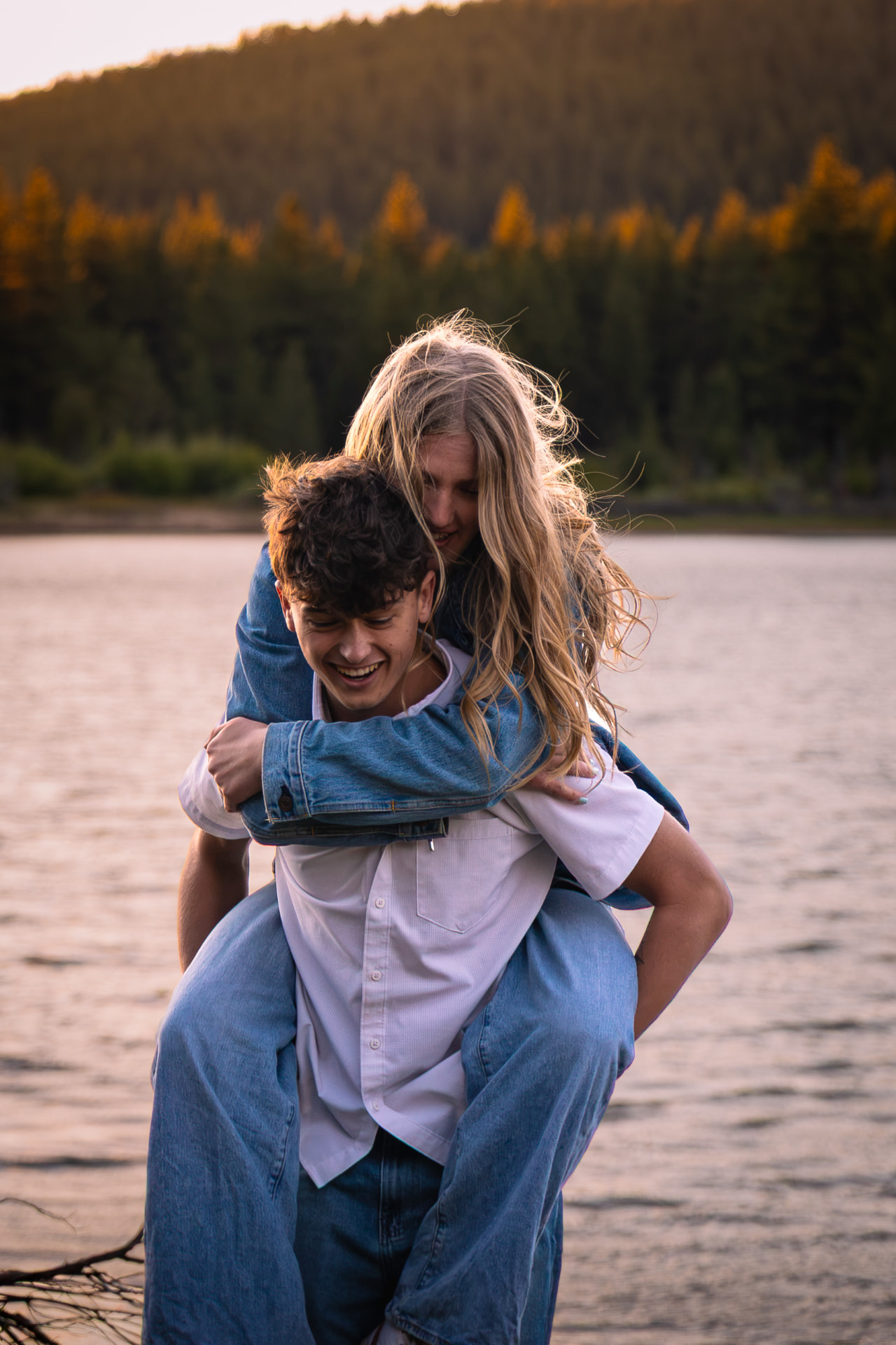 Couple laughing during a piggyback ride beside a mountain lake at golden hour