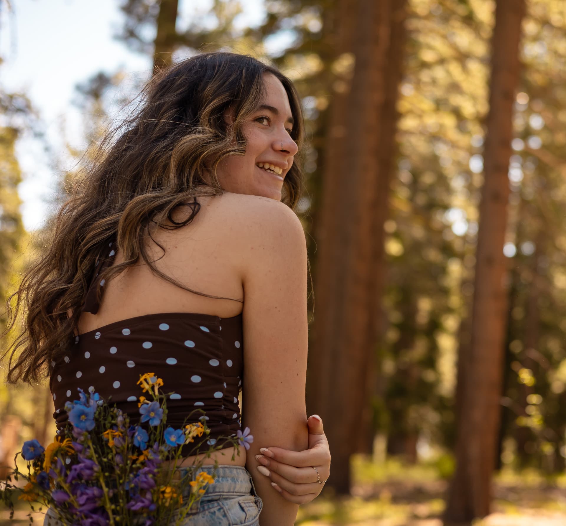 Senior portrait of a young woman glancing back with a bouquet of wildflowers in a pine forest