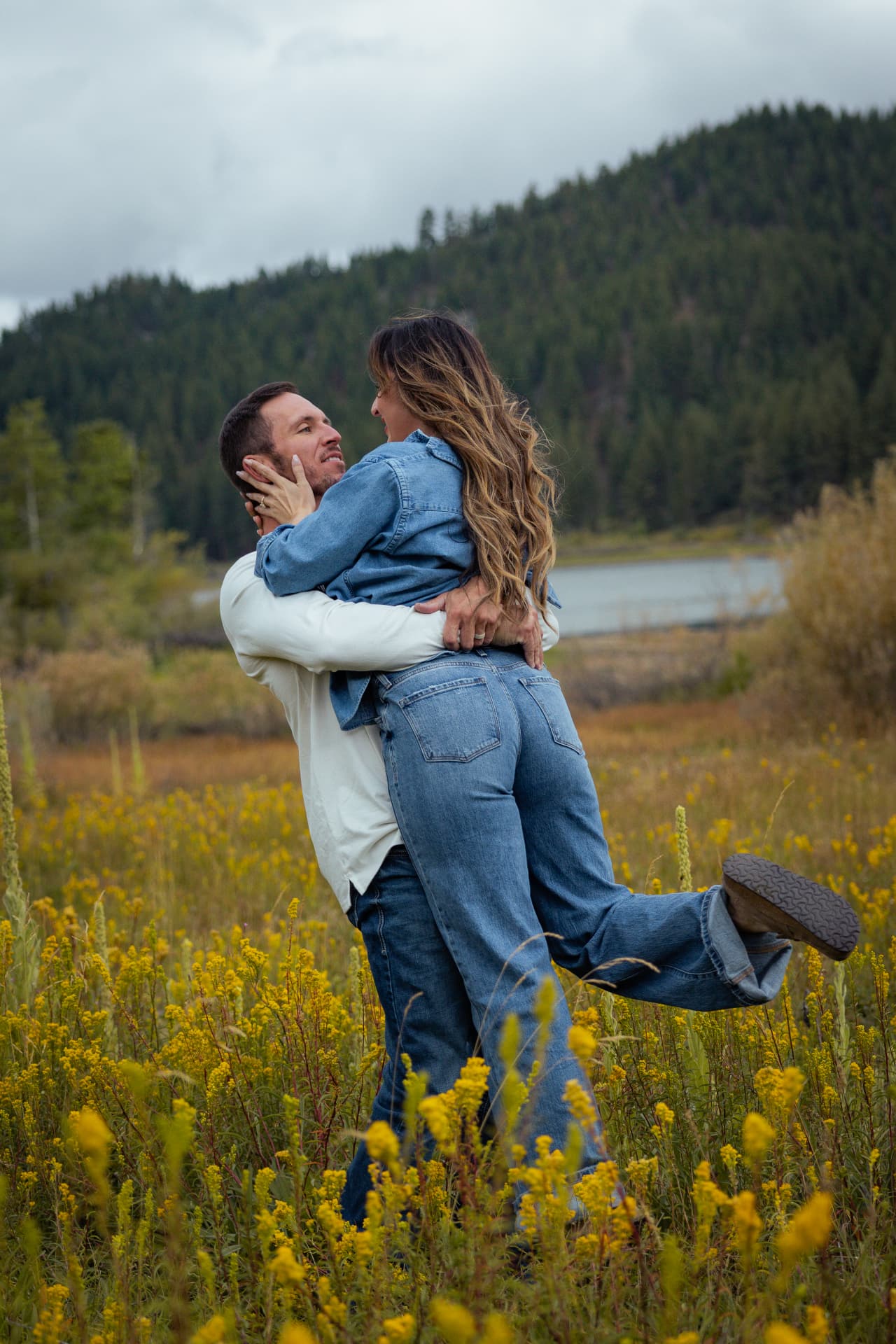 Couple laughing as he lifts her into his arms among golden wildflowers with a mountain lake behind them