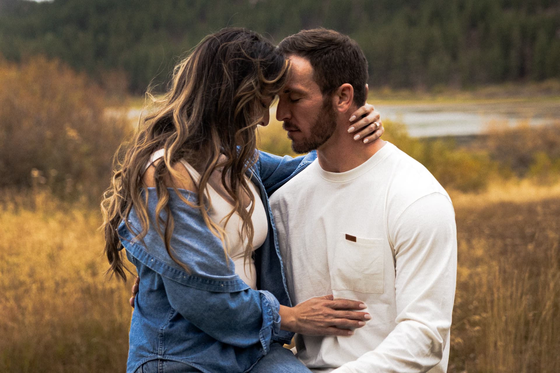 Engaged couple forehead to forehead in a golden autumn meadow, her hand on his neck showing the ring