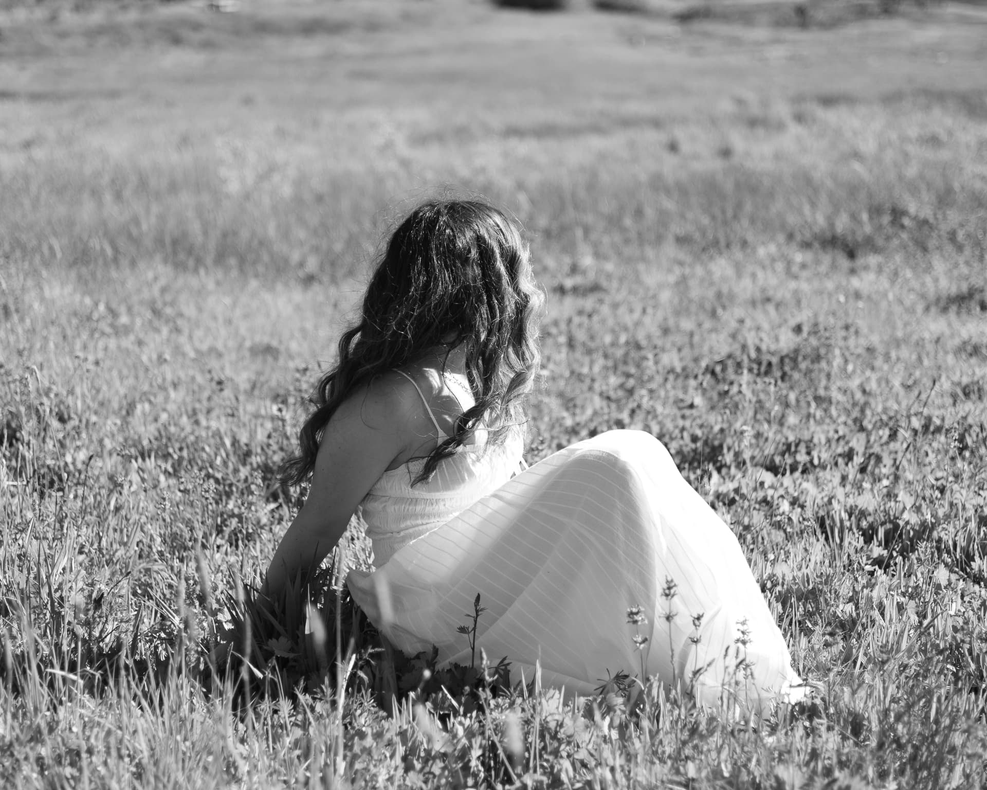 Black and white senior portrait of a young woman in a white dress sitting in a sunlit field