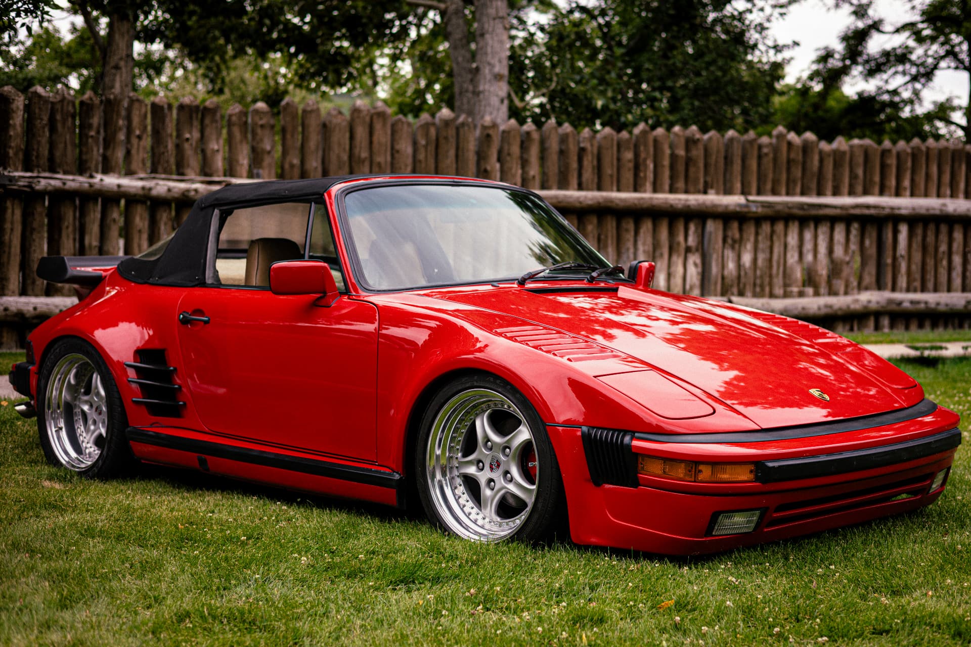 Red slantnose Porsche 911 convertible parked on grass in front of a wooden fence