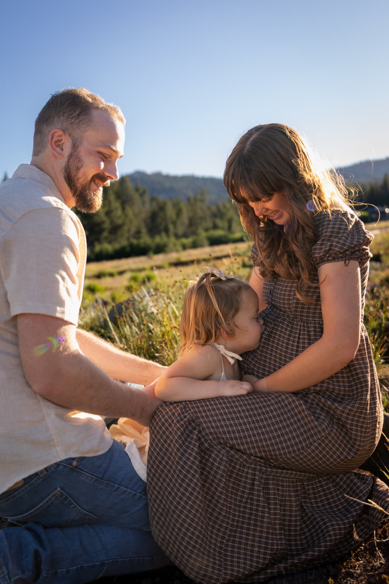 Toddler kissing her expecting mother's belly while dad looks on in a mountain meadow at golden hour