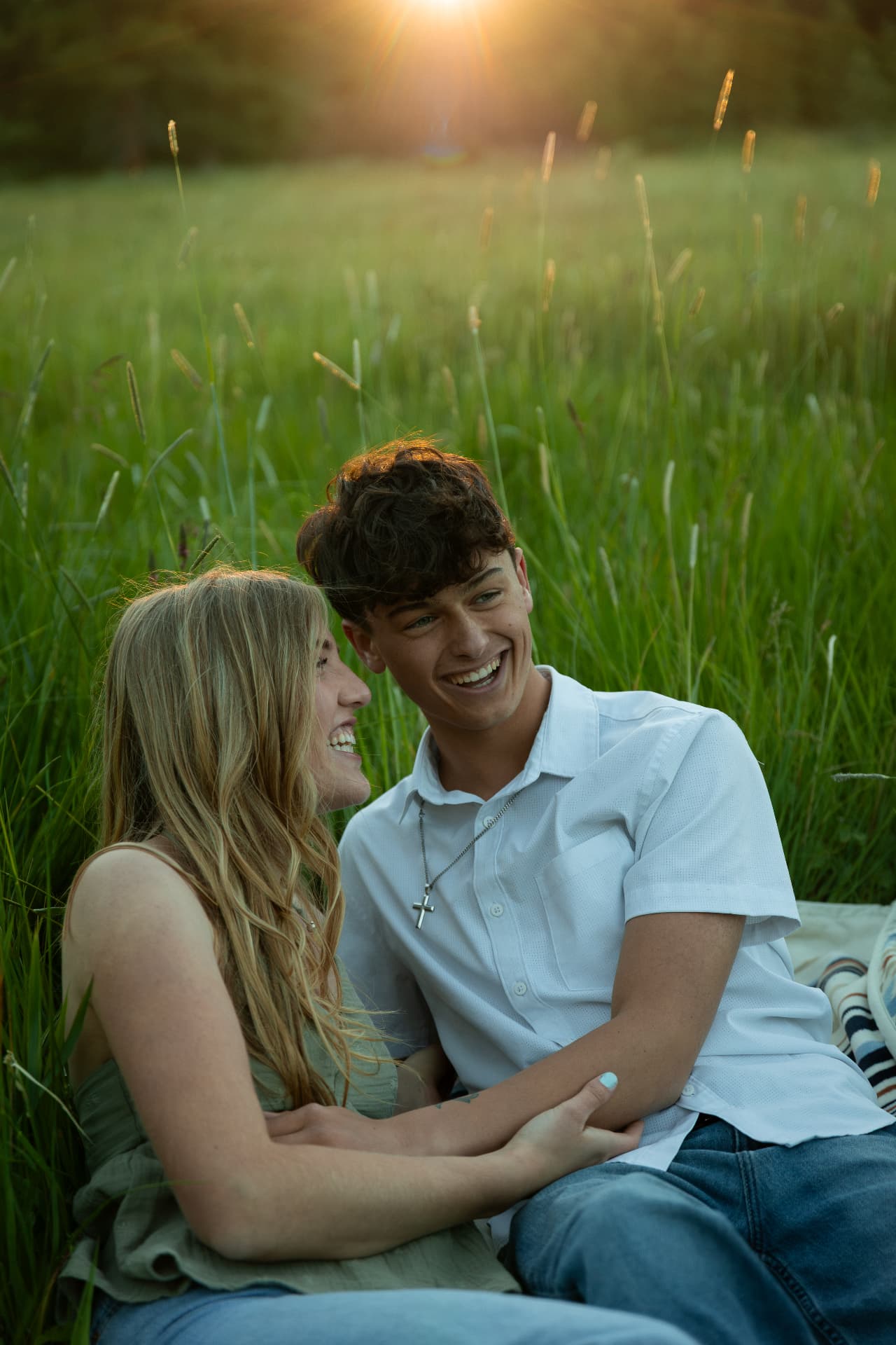 Couple laughing together while sitting in tall sunlit grass at golden hour
