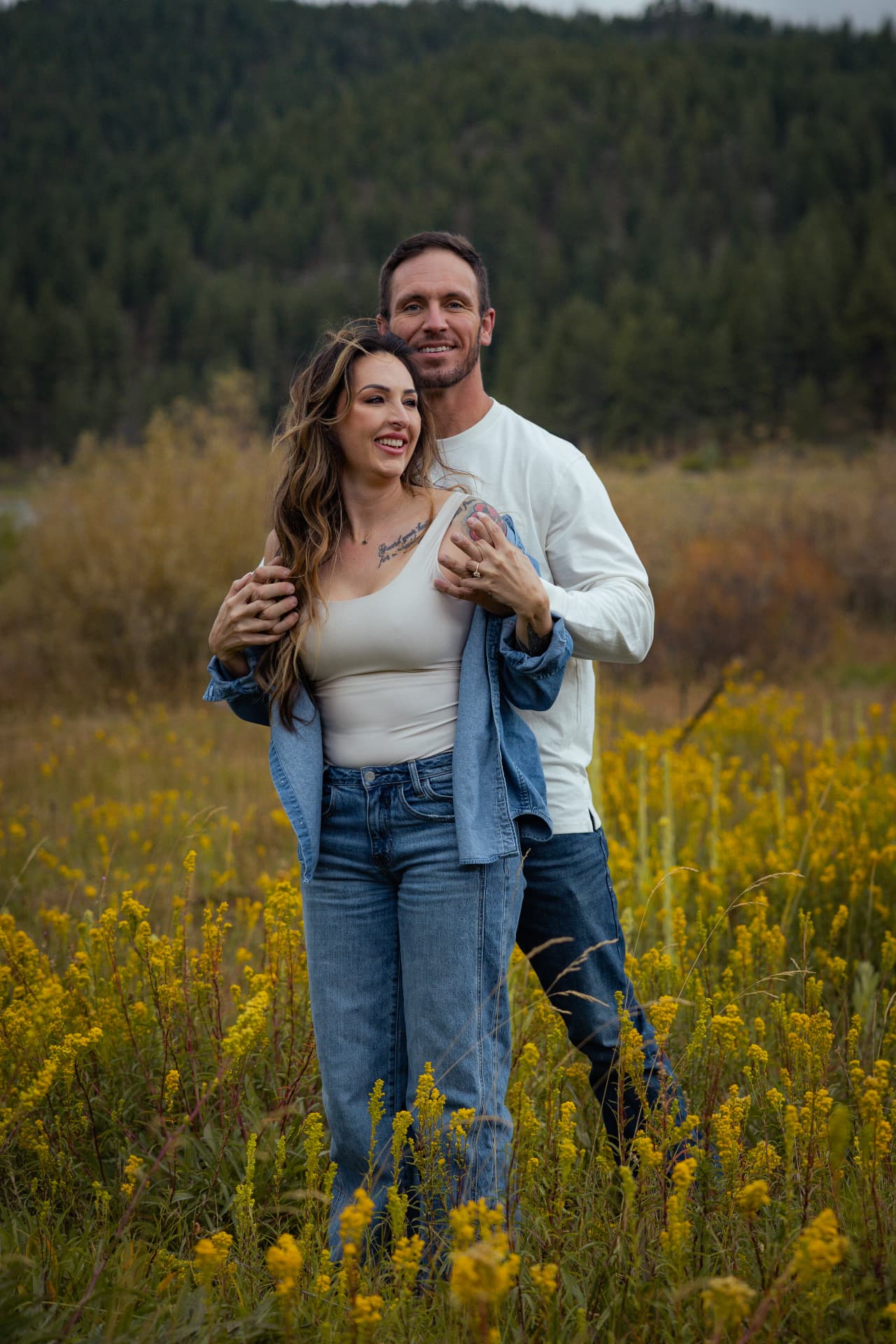 Couple embracing among golden wildflowers with a pine forest behind them
